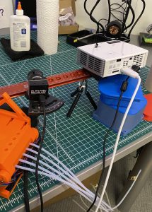 Our scanner setup. The camera and projector are used to take scans, while the dotted board in the upper left is necessary for calibration. The various items under the mount are just there to hold it up; since this image was taken, we’ve mounted the tripod to a more stable wooden board.